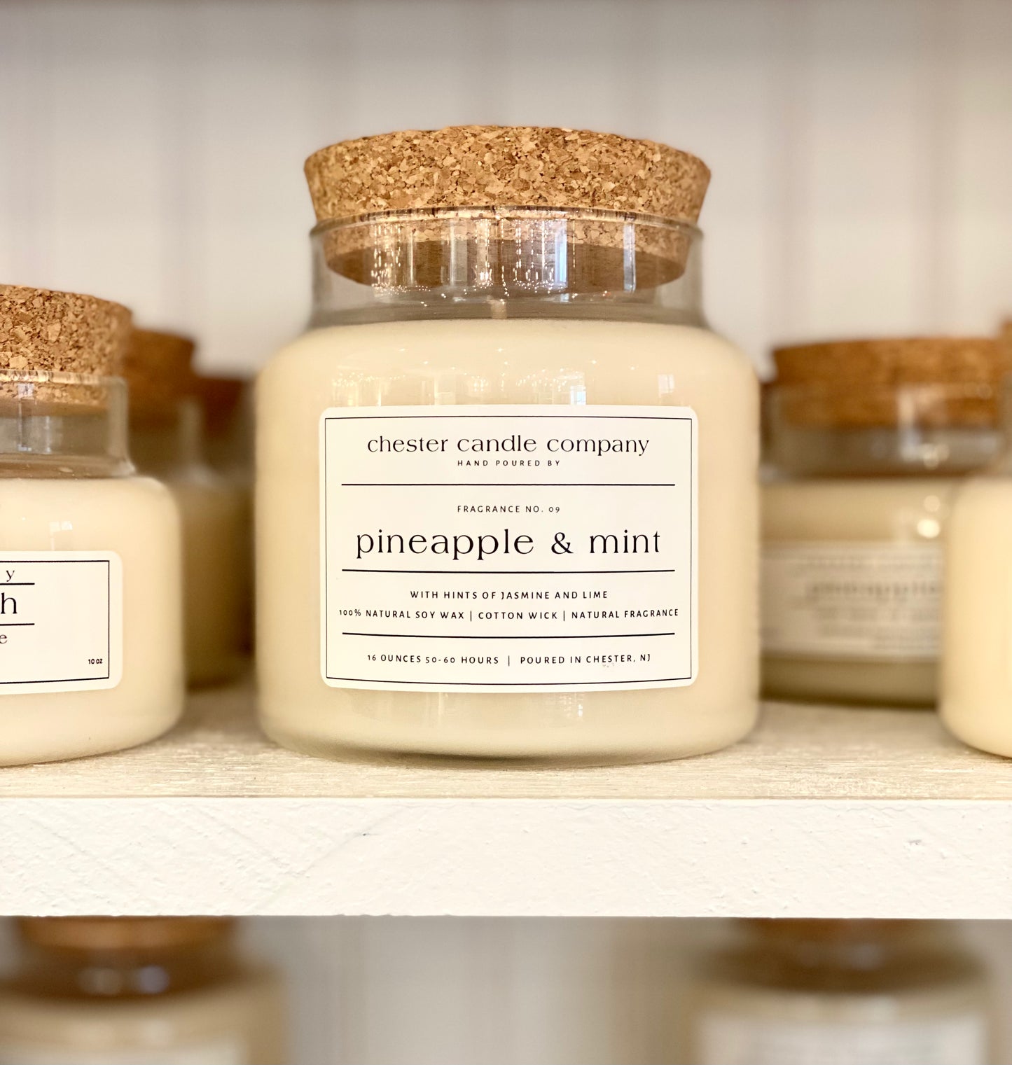 Natural Soy Wax Candle in a Clear Glass Apothecary-Style Jar and a Cork Lid. White Label on the Jar Reads "chester candle company. Fragrance No. 09. pineapple & mint with hints of jasmine and lime. 100% Natural Soy Wax, Cotton Wick, Natural Fragrance. 16 ounces 50-60 hours. Poured in Chester, NJ”