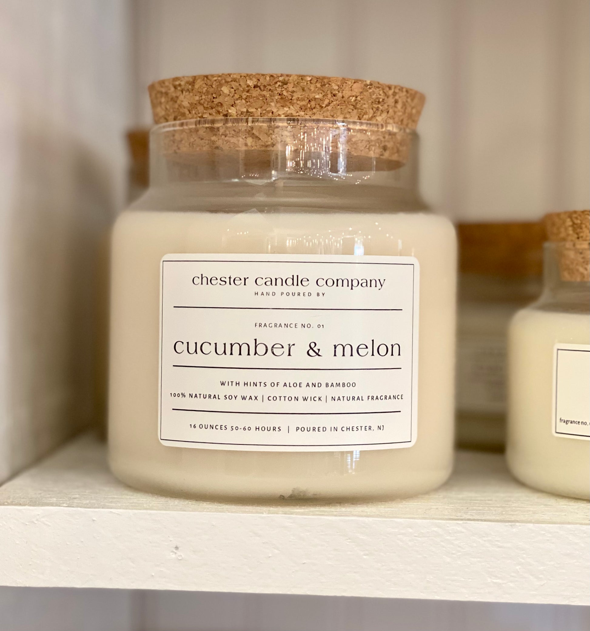 Natural Soy Wax Candle in a Clear Glass Apothecary-Style Jar and a Cork Lid. White Label on the Jar Reads "chester candle company. Fragrance No. 01. cucumber & melon with hints of aloe and bamboo. 100% Natural Soy Wax, Cotton Wick, Natural Fragrance. 16 ounces 50-60 hours. Poured in Chester, NJ”