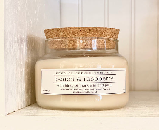 Natural Soy Wax Candle in a Clear Glass Apothecary-Style Jar and a Cork Lid. White Label on the Jar Reads "chester candle company. peach & raspberry with hints of mandarin and plum. 100% American Grown Soy, Cotton Wick, Natural Fragrance. Fragrance No. 02 Hand Poured in Chester, NJ. 10oz