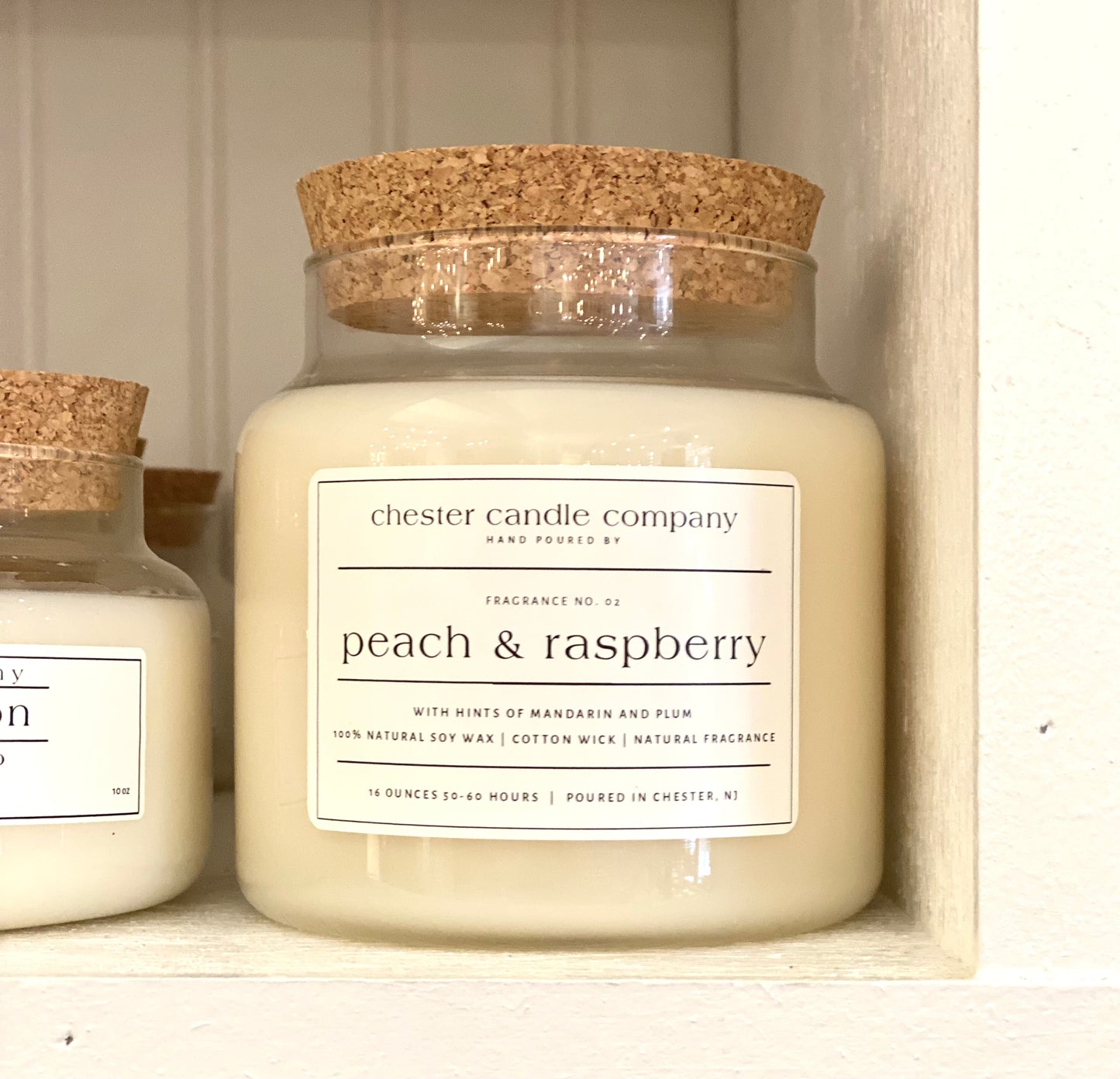 Natural Soy Wax Candle in a Clear Glass Apothecary-Style Jar and a Cork Lid. White Label on the Jar Reads "chester candle company. Fragrance No. 02. peach & raspberry with hints of mandarin and plum. 100% Natural Soy Wax, Cotton Wick, Natural Fragrance. 16 ounces 50-60 hours. Poured in Chester, NJ”