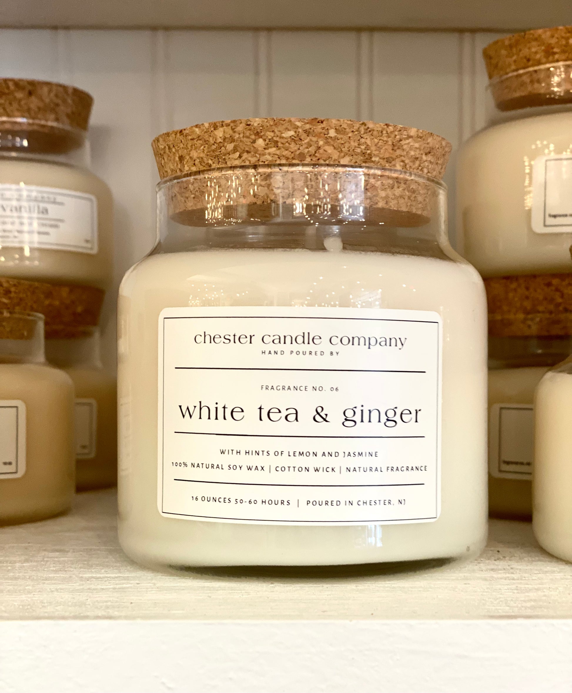 Natural Soy Wax Candle in a Clear Glass Apothecary-Style Jar and a Cork Lid. White Label on the Jar Reads "chester candle company. Fragrance No. 06. white tea & ginger with hints of lemon and jasmine. 100% Natural Soy Wax, Cotton Wick, Natural Fragrance. 16 ounces 50-60 hours. Poured in Chester, NJ”