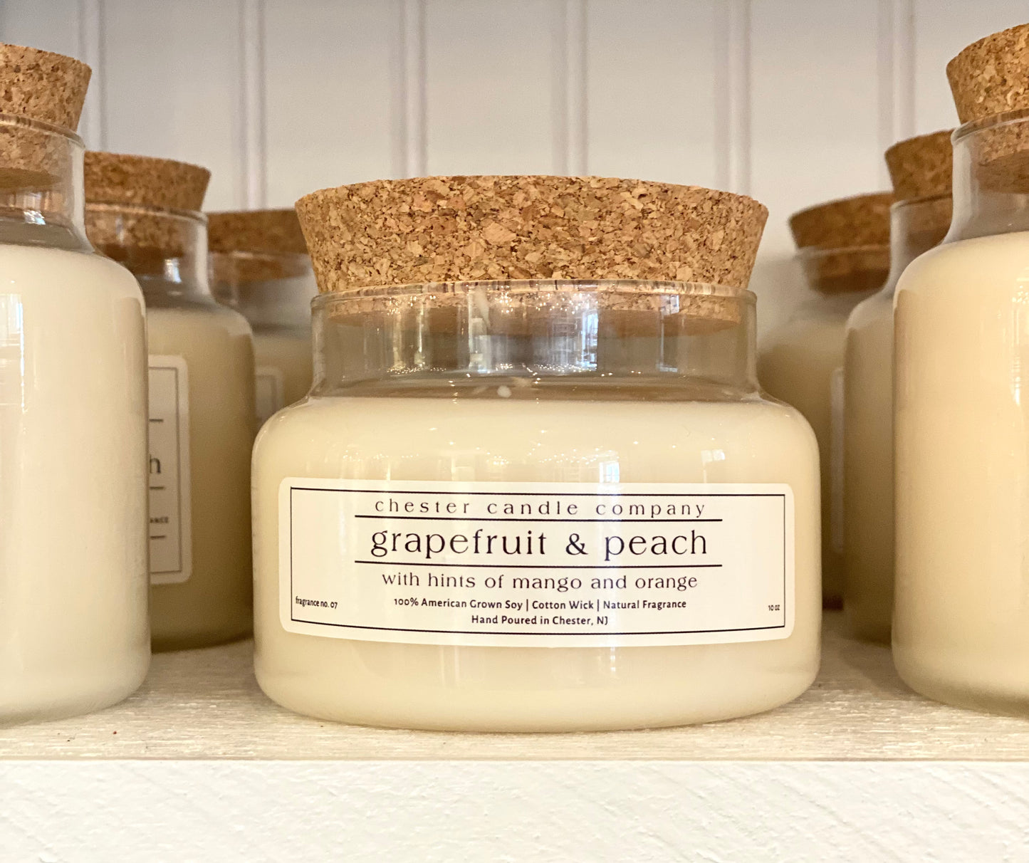 Natural Soy Wax Candle in a Clear Glass Apothecary-Style Jar and a Cork Lid. White Label on the Jar Reads "chester candle company. Grapefruit & peach with hints of mango and orange. 100% American Grown Soy, Cotton Wick, Natural Fragrance. Fragrance No. 07 Hand Poured in Chester, NJ. 10oz”